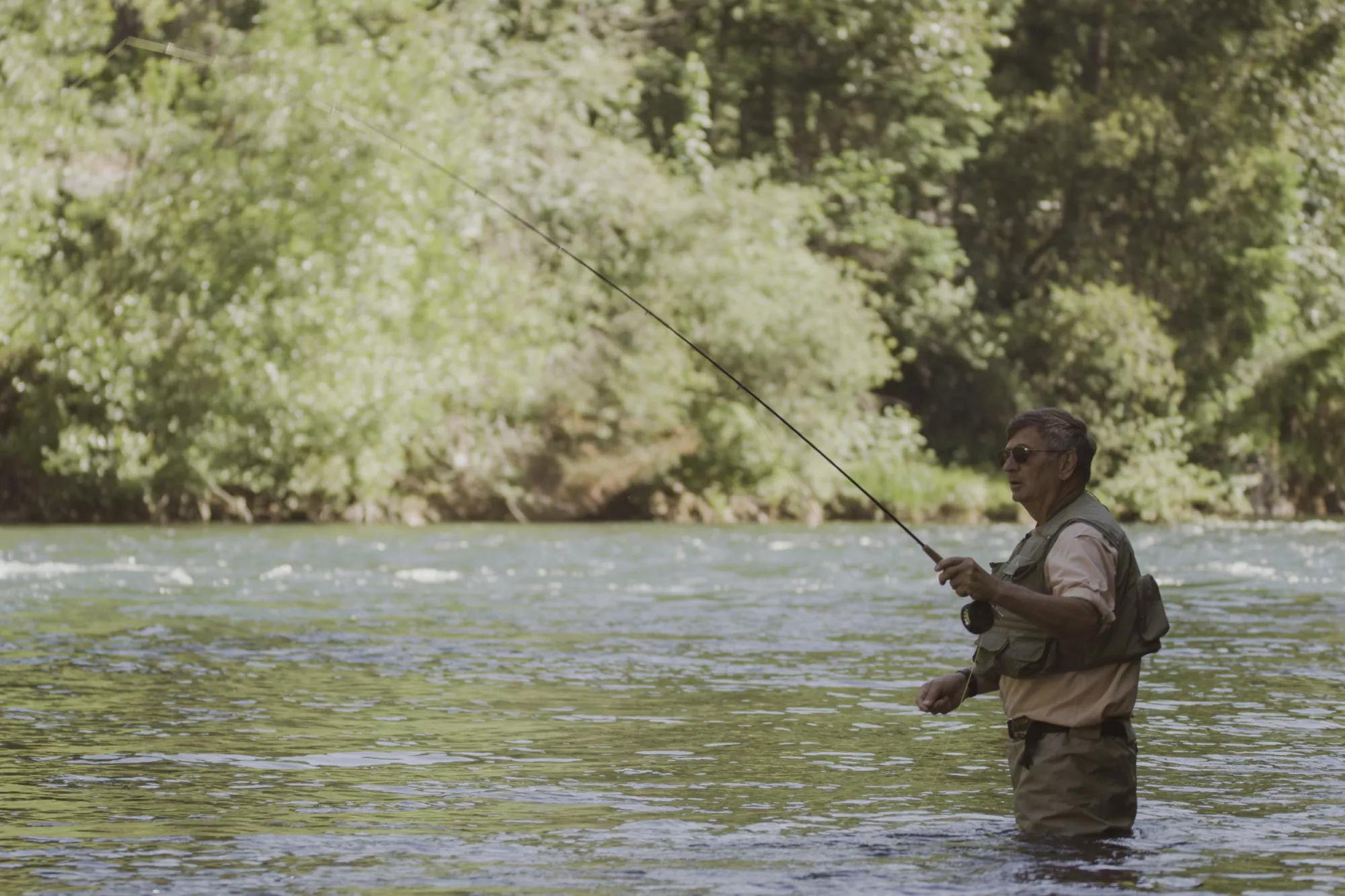 A man fly fishing in a stream wearing waders and sunglasses.