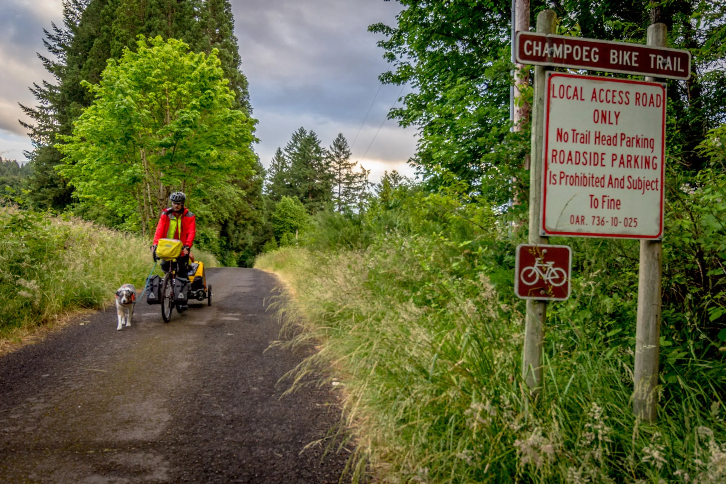 A man rides a bike down the Champoeg Bike Trail pulling a covered wagon with his dog on a leash by his side.