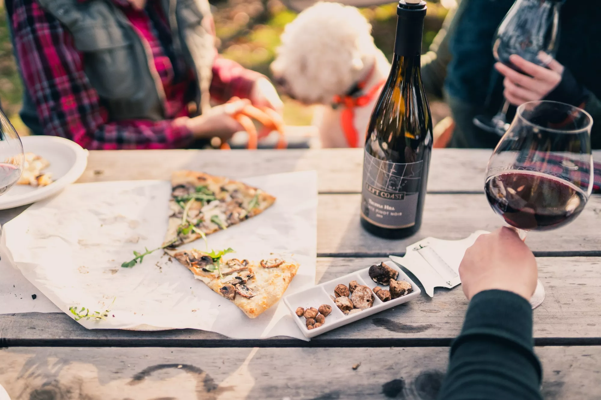 Guests with a dog sit at a table with pizza, truffles, a wine bottle and a full wine glass.