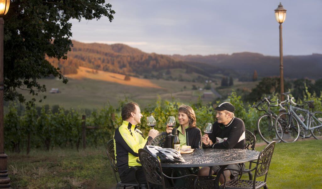 Three people sit at a table drinking wine at Sweet Cheeks Winery, bicycles and rolling hills are in the background