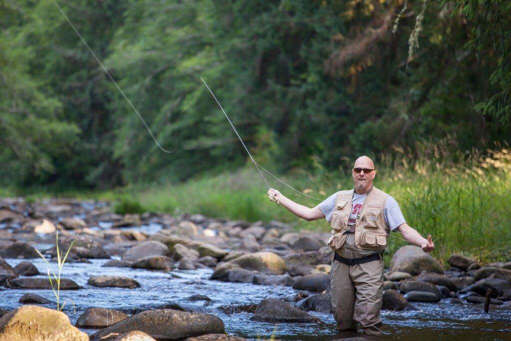 A man standing in a river casting his fly fishing rod while wearing sunglasses.