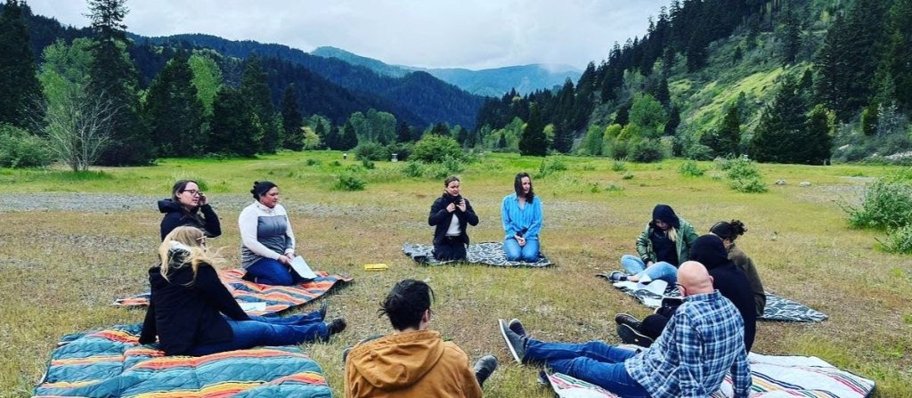 A group of people sitting in a circle in the forest with blankets on the ground. 